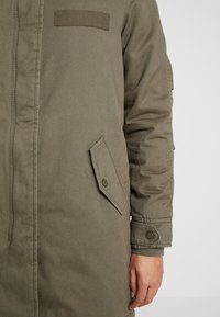 Olivgrüne Jacke aus strukturiertem Stoff, mit einer Knopfleiste vorne, einer diagonalen Tasche und gerippten Bündchen für einen engen Sitz.