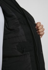 Chaqueta acolchada negra con un bolsillo con cremallera y detalle de cordón en el interior. Textura exterior suave con un diseño ligero.