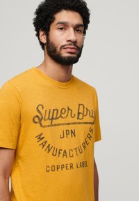 T-shirt di cotone gialla con collo rotondo, caratterizzata da testo stampato in nero: "Super Dry JPN Manufacturers Copper Label." Design casual e strutturato.