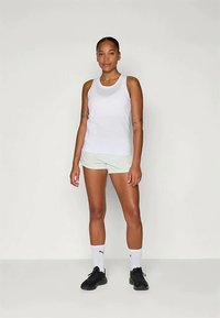 Frau steht in weißem Tanktop, hellen Shorts, weißen Puma-Socken und schwarzen Turnschuhen vor einem schlichten hellen Hintergrund.
