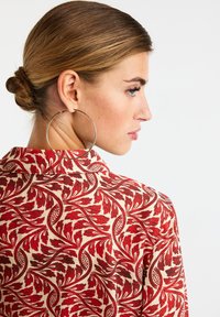 Košeľa s červeným kvetinovým vzorom a golierom, s krémovým pozadím. Modelka má účes do drdolu a veľké kruhové náušnice.