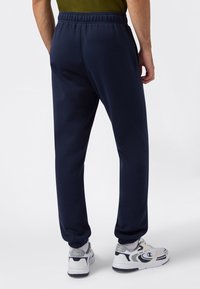 Mężczyzna w granatowych dresach z elastycznymi mankietami oraz białych, szarych i czarnych sneakersach, stojący na jednolitym tle.