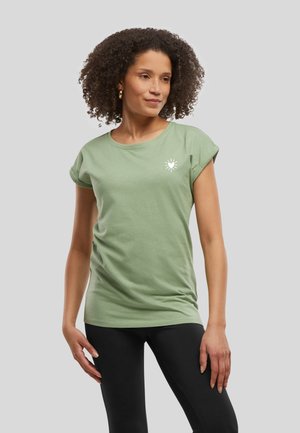 Femme aux cheveux bouclés portant un t-shirt vert clair à manches courtes avec un petit motif de cœur blanc et des leggings noirs, debout devant un fond uni.