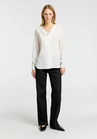 Hvid langærmet bluse med V-hals og subtil glans, parret med sorte lige bukser og sorte spidse flade sko.