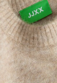 Beżowy sweter dzianinowy z prążkowanym kołnierzem. Posiada wyraźną zieloną metkę z białym napisem "JJXX". Miękka, teksturowana tkanina.