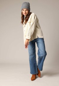 Witte corduroy overhemdjas met grote knopen, gecombineerd met blauwe wijde jeans en bruine suède enkellaarsjes. Grijze gebreide beanie op het hoofd.