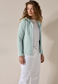Felpa blu chiaro con zip, in tessuto testurizzato, con coulisse e tasche laterali, abbinata a pantaloni bianchi a gamba larga.
