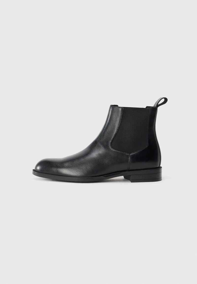 Botín negro de cuero hasta el tobillo con paneles elásticos a los lados, punta redondeada y lengüeta. Textura suave, suela plana, diseño minimalista.
