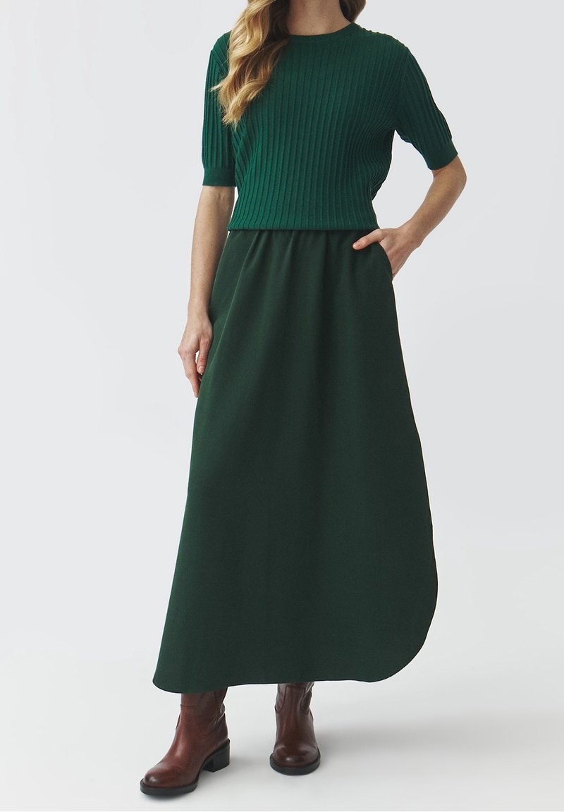 Groene geribbelde gebreide top met korte mouwen, gecombineerd met een lange, vloeiende groene rok, gedragen met bruine enkelboots met een lage blokhak.