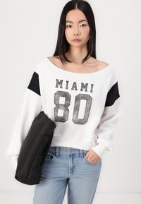 Sudadera blanca con detalles negros, que presenta un gráfico de "MIAMI 80". El modelo sostiene un cojín gris oscuro y lleva unos jeans de color azul claro.