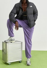 Czarna puchowa kurtka, fioletowe dresy i szare trampki na srebrnej walizce na kółkach. Materiał o fakturze, zapięcie na zamek i dopasowany krój.