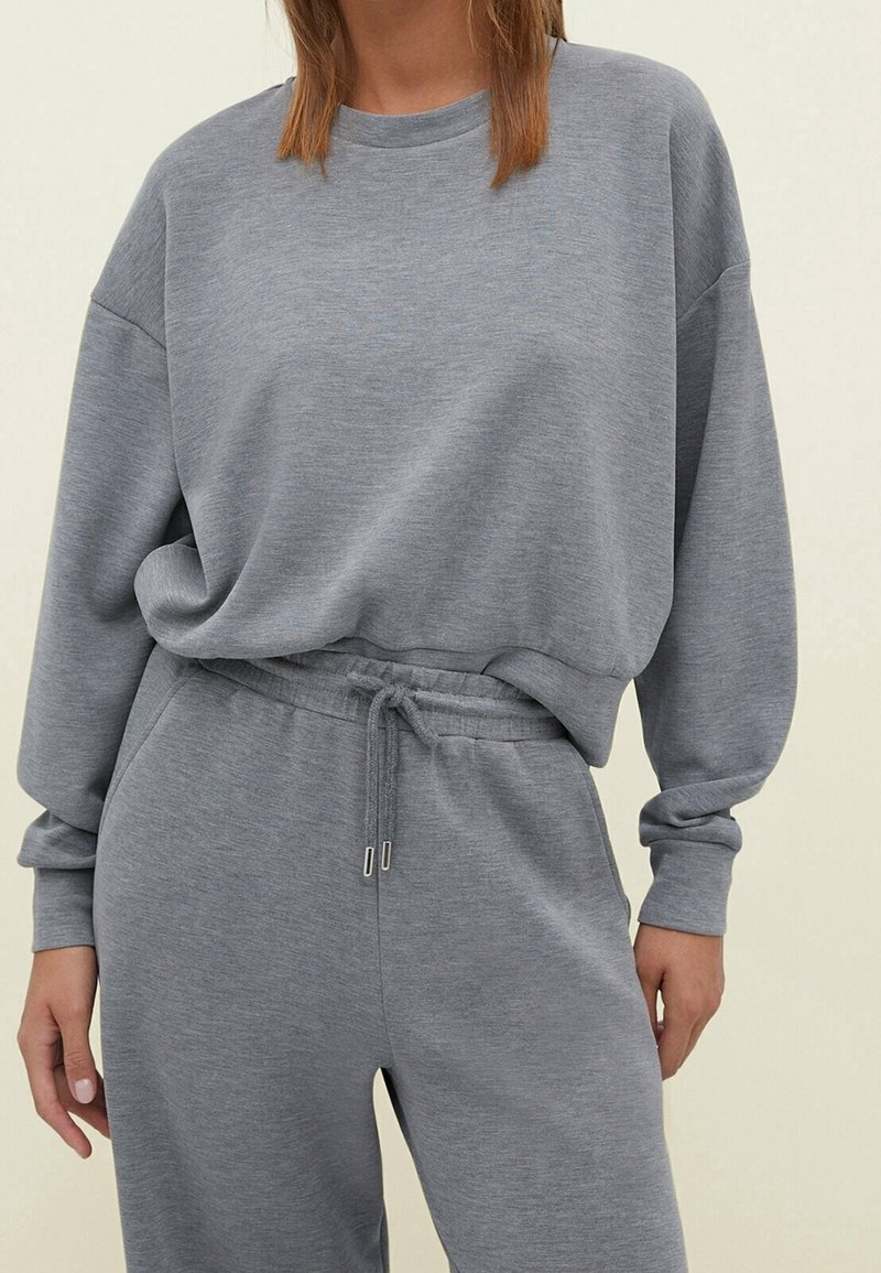 Felpa grigia e pantaloni coordinati realizzati in un morbido tessuto testurizzato. La felpa ha una vestibilità ampia e polsini a coste. I pantaloni presentano un girovita con cordino.
