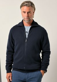 Giacca con zip blu navy realizzata in tessuto morbido, caratterizzata da collo alto, polsini a costine e una cerniera, abbinata a una maglietta azzurro chiaro sotto.