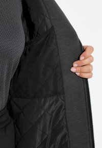 Czarna kurtka od wewnątrz z pikowaną podszewką, siatkową kieszenią i teksturowanym szarym materiałem. Zamek błyskawiczny po boku, ręka trzymająca brzeg.
