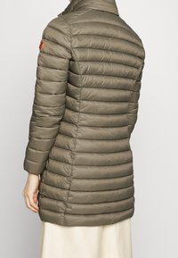 Doudoune kaki avec des coutures horizontales, col haut et poches latérales. Elle arbore un patch logo sur l'épaule gauche. Tissu lisse et brillant.