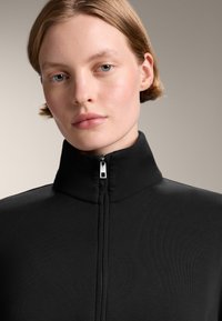 Černý zipovací top s vysokým límcem vyrobený z rib rovnajícího se materiálu; má stříbrný zip, hladký povrch a přiléhavý střih.