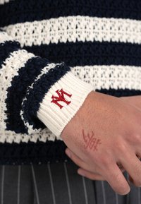 Muñeca con puño blanco con logo rojo de NY en manga de punto a rayas azul marino y blanco, mostrando un tatuaje de tinta roja en la mano.