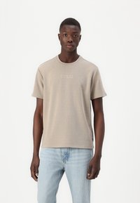 Beige tričko s krátkým rukávem, s vroubkovanou texturou a kulatým výstřihem, na hrudi vyšívané logo "GUESS". Nosí se s světle modrými džíny.
