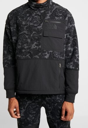 Mężczyzna ubrany w czarną i ciemnoszarą polarową bluzę z nadrukiem, z kieszeniami na piersi i bokach zapinanymi na zamek, z logo The North Face na kieszeni na piersi.