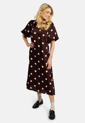 Lyshåret kvinde iført en mørkebrun midi-kjole med lyserøde polkaprikker, beige sokker og sorte loafers, smilende med hånden på hoften.