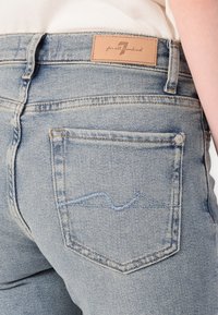 Jeans din denim de culoare albastru deschis au o etichetă din piele la talie în spate, cu un model de cusătură curbat pe buzunarul din dreapta.