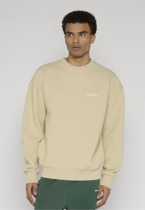 Mand iført beige tekstureret sweatshirt og mørkegrønne bukser, står foran en ensfarvet grå baggrund og ser mod venstre.