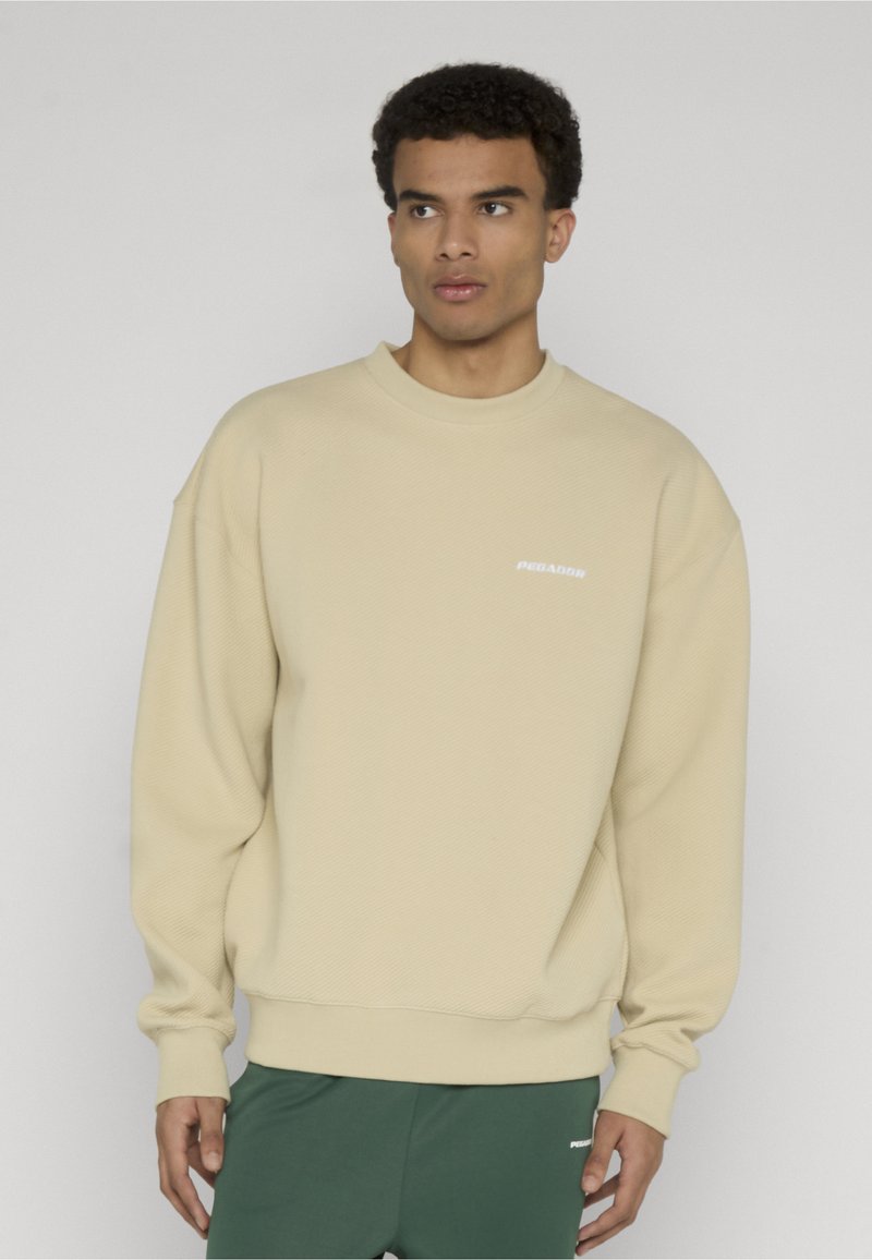 Hombre con sudadera beige texturizada y pantalones verde oscuro, de pie frente a un fondo gris liso, mirando hacia su izquierda.