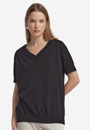 Kvinde iført en løs sort V-hals kortærmet top og sølv øreringe i hoop-stil, stående mod en ensfarvet hvid baggrund.