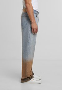 Vyras vilkintis šviesiai mėlynus džinsus su rudos spalvos gradientu apačioje, baltus sportbačius ir baltą marškinį prieš neutralų foną.