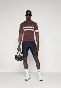 Tamsiai ruda dviračių džersio marškinėliai su baltomis juostelėmis, trumpomis rankovėmis ir priekyje užtrauktuku. Dėvima su juodais šortais ir baltais kojinėmis. Šalmas laikomas rankoje.