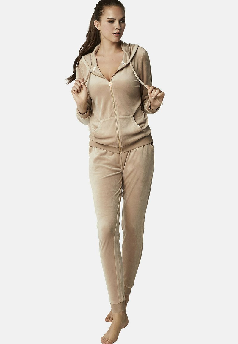 Ensemble de survêtement en velours beige comprenant une veste à capuche zippée et un pantalon ajusté avec des chevilles élastiquées et des poches latérales. Texture lisse, coupe décontractée.
