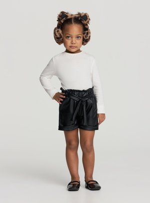 Bambino con maglia bianca a maniche lunghe, pantaloncini corti in ecopelle nera con cintura annodata in vita e paraorecchie con stampa animalier, in piedi con sicurezza con una mano sui fianchi.
