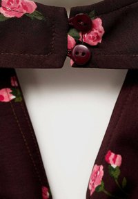 Cvjetna bluza od burgundske tkanine, s uzorkom ružičastih ruža. Dva okrugla maroon gumba s po dva otvora svaki u blizini ovratnika.