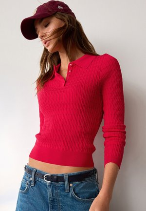 Donna che indossa un maglione polo rosso a maniche lunghe con trama, jeans blu con cintura nera e un cappellino da baseball rosso scuro, su sfondo bianco.