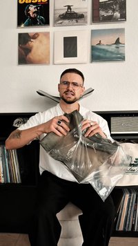 Zaļgani brūni bikses caurspīdīgā plastmasas maisiņā, ar taisnu kāju dizainu. Vīrietis tur bikses, apkārt ir ieraksti un skaļrunis.