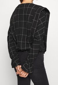 Svart rutigt blus i strukturerad tyg, långa ärmar med samlade manschetter, och rundad halsringning, matchad med svarta byxor.