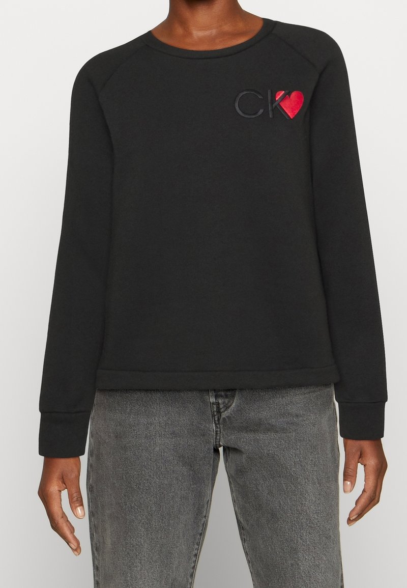 Felpa nera con scollatura rotonda e maniche raglan. Presenta un accento a forma di cuore rosso combinato con il logo "CK" in ricamo nero.