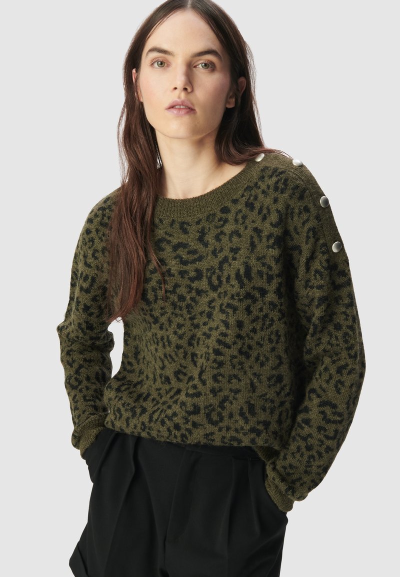 Vrouw met lang bruin haar, gekleed in een olijfgroene en zwarte luipaardprinttrui met zilveren knopen op de schouder, met handen in de zakken van zwarte broek.