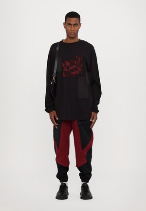 Schwarzer, übergroßer Sweatshirt mit rotem Grafik-Patch, kombiniert mit roten und schwarzen strukturierten Hosen mit eckigen Ausschnitten. Schwarze Sneakers und eine Umhängetasche.