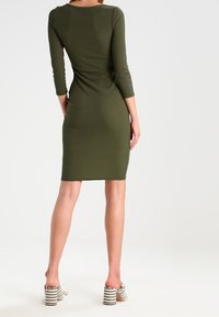 Olivově zelené žebrované šaty s tříčtvrtečními rukávy a přiléhavým střihem, doplněné béžovými a černými pruhovanými sandály na blokovém podpatku.