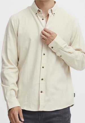 Person wearing a long-sleeve beige button-up shirt, partially buttoned, with one hand adjusting the collar near the neck.