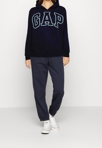 Mörkblå huvtröja med "GAP"-logotyp i ljusblått, utrustad med dragsko och ribbade ärmslut. Matchas med mörkgrå joggerbyxor och vita sneakers.