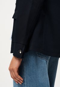 Χέρι χαλαρό στο πλάι, φοράει σκούρο σακάκι με κουμπί μανσέτας και ανοιχτό μπλε τζιν με ορατή ραφή στην πίσω τσέπη.