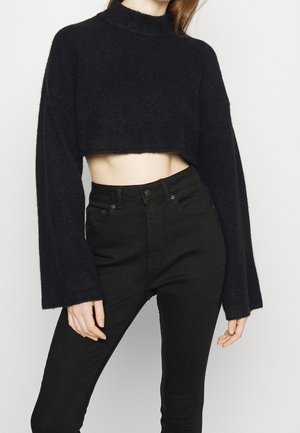 Femme portant un pull court noir à manches longues et un jean noir taille haute, debout devant un fond uni.
