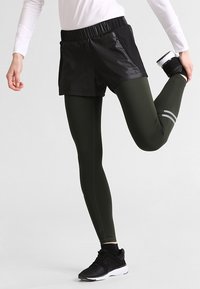 Svarta löparshorts över mörkgröna leggings, med resårmidja och dragkedjefickor, kombinerat med svarta sportskor.