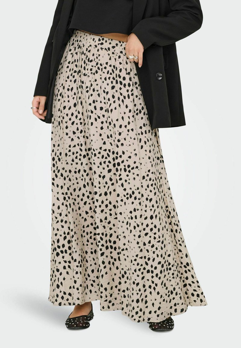 Donna che indossa una lunga gonna beige con macchie nere irregolari, un top nero corto, un blazer nero e ballerine a pois neri.