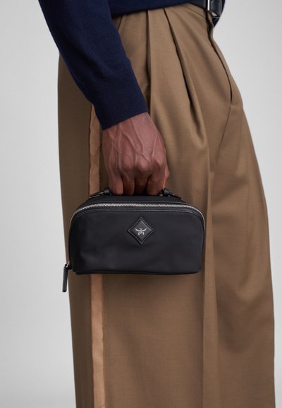 Necessaire de nylon preto com fecho éclair, logotipo em formato de diamante e alças duplas; segurada por uma pessoa ao lado de calças beige plissadas.