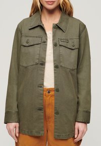 Olijfgroen knopenjasje met vier borstzakken, een kraag en lange mouwen. Draag het over een beige top en oranje broek.