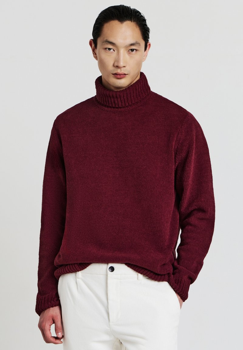 Junger Mann in einem burgunderfarbenen Rollkragenpullover und weißen Hosen, der vor einem einfarbigen hellgrauen Hintergrund steht.