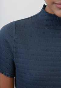 Tmavě modrý žebrovaný top s vysokým výstřihem a krátkými rukávy. Tkanina vypadá texturovaně s jemnými vzory a má čistý lem.